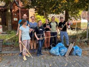 Eine fröhliche Gruppe Erwachsener mit Holzgreifzangen steht auf einem Gehweg unter Bäumen neben gefüllten blauen Müllsäcken und Holzlatten.