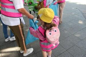Ein Kind mit gelbem Hut und pinker Weste sammelt mit einer Erzieherin Müll in einen blauen Sack auf einem Gehweg.