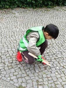 Ein Junge in grüner World-Cleanup-Day-Weste hockt mit Holzgreifzange auf Kopfsteinpflaster und greift nach einem kleinen Fundstück.