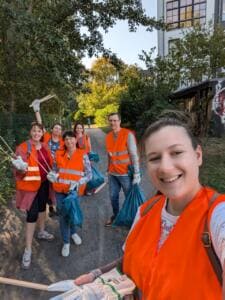 Selfie einer Frau in orangefarbener Warnweste, hinter ihr winkt eine Gruppe in Warnwesten mit Greifzangen und Säcken auf einem Weg.
