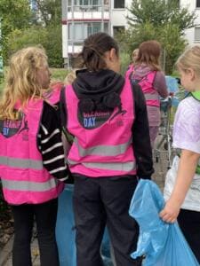Schülerinnen in pinken World-Cleanup-Day-Westen stehen von hinten fotografiert mit blauen Müllsäcken auf einem Gehweg.