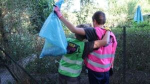 Zwei Kinder in World-Cleanup-Day-Westen stehen von hinten fotografiert an einem Zaun, eines hält einen vollen blauen Müllsack hoch.