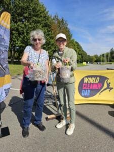 Zwei ältere Frauen zeigen lachend Gläser voller gesammelter Zigarettenstummel vor einem gelben World-Cleanup-Day-Banner.