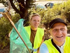 Selfie eines Mannes mit Kappe und einer Frau in gelben Warnwesten mit Greifzange und grünem Müllsack vor Büschen.