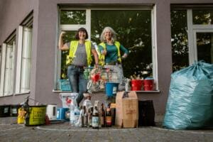 Zwei Frauen in gelben Warnwesten posieren vor einem Fenster hinter vielen Fundstücken wie Flaschen, Eimern, einem Einkaufswagen und einem Trash-Hero-Eimer.