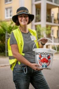Eine lächelnde Frau mit Hut und gelber Warnweste hält einen Eimer mit der Aufschrift Bachgotti's Crabbing Bucket.