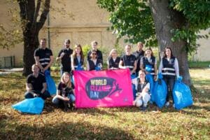 Eine Gruppe in schwarzen Warnwesten posiert mit einem Hund hinter einer pinken World-Cleanup-Day-Fahne und gefüllten blauen Müllsäcken auf herbstlichem Laub.