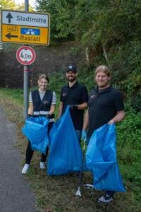 Drei Erwachsene in schwarzer Kleidung halten volle blaue Müllsäcke am Straßenrand neben einem Wegweiser nach Rastatt.