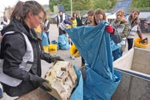 Eine Frau in weißer Warnweste hebt einen verschmutzten Kanister in einen Container, daneben sortieren weitere Helferinnen Müll in blaue Säcke.