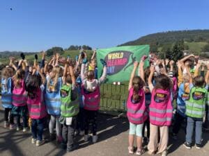 Viele Kinder in bunten World-Cleanup-Day-Westen recken von hinten fotografiert die Arme vor einer grünen World-Cleanup-Day-Fahne, im Hintergrund Weinberge.
