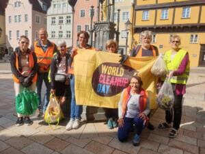 Eine Gruppe älterer Erwachsener in Warnwesten hält in der Altstadt eine gelbe World-Cleanup-Day-Fahne und Beutel mit gesammeltem Müll.