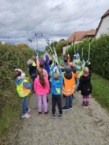 Eine Gruppe Kinder in Warnwesten steht von hinten fotografiert auf einem Schotterweg zwischen Hecken und reckt die Greifzangen in die Luft.