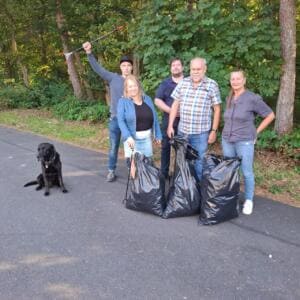 Fünf Erwachsene stehen am Waldweg hinter drei vollen schwarzen Müllsäcken, ein Mann reckt eine Greifzange hoch, daneben sitzt ein schwarzer Hund.