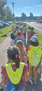 Eine lange Reihe Grundschulkinder mit Rucksäcken und Warnwesten läuft einen Gehweg an einer Straße entlang, von hinten fotografiert.