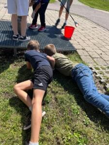 Zwei Jungen liegen bäuchlings im Gras und spähen unter einen Gitterrost; daneben stehen weitere Kinder mit Greifzange und rotem Eimer.