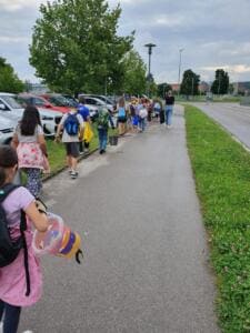 Grundschulkinder mit Eimern und Rucksäcken gehen hintereinander einen Gehweg neben parkenden Autos entlang.