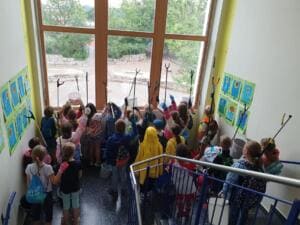 Viele Grundschulkinder mit Eimern und Greifzangen stehen von hinten fotografiert in einem Treppenhaus vor einem großen Fenster und heben die Zangen.
