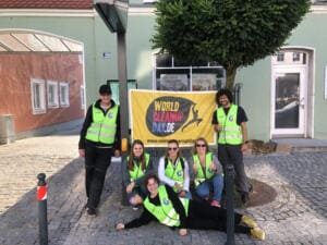 Sechs Erwachsene in gelben Warnwesten posieren lachend vor einem gelben World-Cleanup-Day-Banner, eine Frau liegt vorne auf dem Pflaster.
