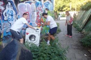 Zwei junge Männer ziehen eine weggeworfene Waschmaschine aus dem Gebüsch neben einer mit Graffiti besprühten Wand.
