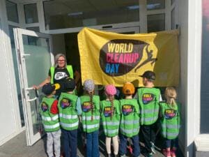 Kindergartenkinder in grünen World-Cleanup-Day-Westen stehen von hinten fotografiert mit einer Erzieherin vor einer gelben World-Cleanup-Day-Fahne.