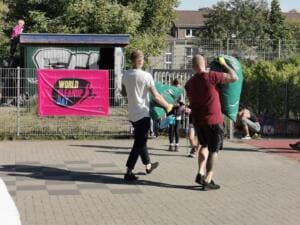 Zwei Männer tragen gefüllte grüne Müllsäcke über einen gepflasterten Platz, im Hintergrund eine pinke World-Cleanup-Day-Fahne an einem Zaun.