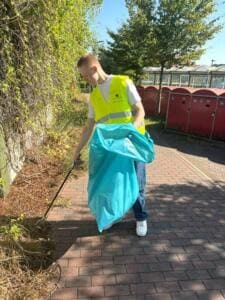 Ein junger Mann in gelber Warnweste sammelt mit Greifzange Müll in einen türkisfarbenen Sack neben roten Fahrradboxen.