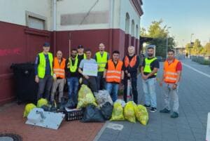 Eine Gruppe Männer in gelben und orangefarbenen Warnwesten steht am Bahnhof neben vielen gefüllten gelben und schwarzen Säcken und einem alten Waschbecken.