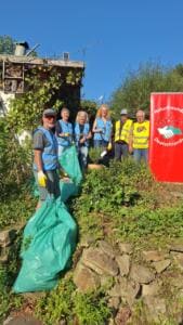 Sechs Erwachsene in blauen und gelben Warnwesten stehen an einem bewachsenen Hang neben gefüllten grünen Müllsäcken und einem roten Banner der NaturFreunde Deutschlands.