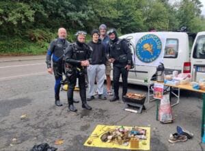 Vier Taucher in Neoprenanzügen und ein Jugendlicher stehen vor einem Transporter mit dem Banner des Tauchclubs Octopus Siegen; davor liegt auf einer gelben Plane der aus dem Wasser geborgene Müll.