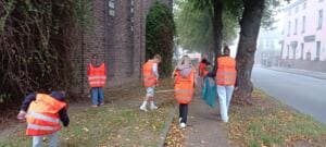 Kinder und Jugendliche in orangefarbenen Warnwesten sammeln Müll auf einem Grünstreifen unter Bäumen entlang einer Straße, von hinten fotografiert.
