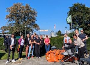 Eine Gruppe Erwachsener steht vor einer Hecke neben vielen orangefarbenen Müllsäcken und einem Einkaufswagen, ein Mann sitzt im Wagen.