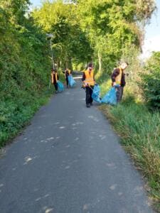 Mehrere Erwachsene in orangefarbenen Warnwesten sammeln mit Greifzangen und blauen Säcken Müll entlang eines grünen Radwegs.