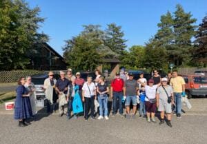 Eine große Gruppe Erwachsener mit Säcken und Greifzangen steht auf einem Parkplatz unter blauem Himmel.