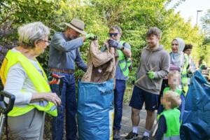 Eine Gruppe aus Erwachsenen und Kindern in Warnwesten steht um einen großen blauen Sack, zwei Männer ziehen eine alte Jacke heraus; zwei Kindergesichter sind unkenntlich gemacht.
