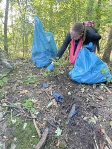 Eine Frau mit pinkem Schal und Handschuhen sammelt Müll vom Waldboden in einen blauen Sack, daneben ein zweiter Sack zwischen Büschen.