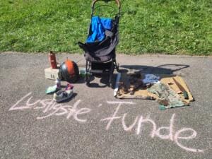 Unter der Kreideaufschrift Lustigste Funde liegen auf dem Asphalt ein Kinderwagen, ein Motorradhelm, ein Schuh, eine Flasche und rostige Metallteile.