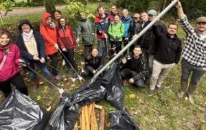 Eine große Gruppe Erwachsener in Regenjacken steht im Kreis und hält Greifzangen und eine Stange über gefüllte schwarze Müllsäcke und Holzlatten.