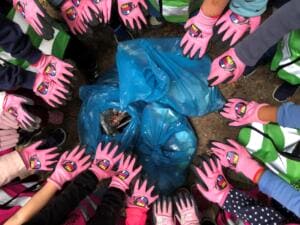 Von oben fotografiert: Viele Kinderhände in pinken World-Cleanup-Day-Handschuhen liegen im Kreis um gefüllte blaue Müllsäcke.