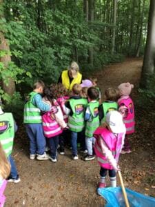 Kindergartenkinder in grünen und pinken World-Cleanup-Day-Westen stehen dicht zusammen mit einer Erzieherin auf einem Waldweg.