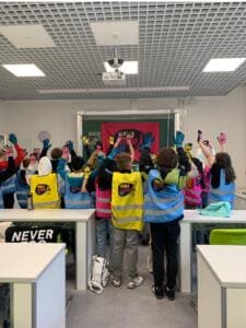 Eine Schulklasse in World-Cleanup-Day-Warnwesten steht von hinten im Klassenzimmer und hebt die Hände mit bunten Arbeitshandschuhen; an der Tafel hängt eine World-Cleanup-Day-Fahne.