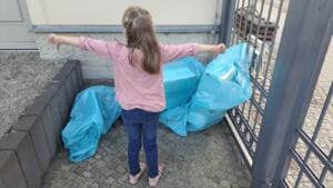Ein Mädchen steht von hinten fotografiert mit ausgebreiteten Armen vor mehreren gefüllten hellblauen Müllsäcken an einem Tor.