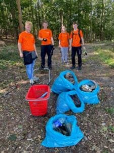 Vier Jugendliche in orangefarbenen T-Shirts stehen auf einem Waldweg hinter gefüllten blauen Säcken und einem roten Einkaufskorb.