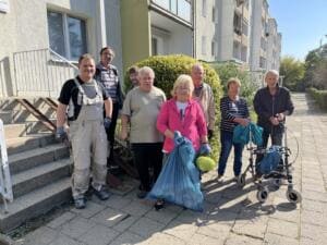 Eine Gruppe älterer Nachbarinnen und Nachbarn steht vor einem Wohnblock mit Müllsäcken und Handschuhen, eine Frau mit Rollator.