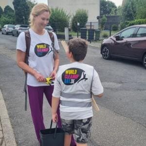 Eine Frau im World-Cleanup-Day-T-Shirt reicht einem Jungen in World-Cleanup-Day-Warnweste Arbeitshandschuhe; der Junge hält einen Eimer und eine Holzlatte.