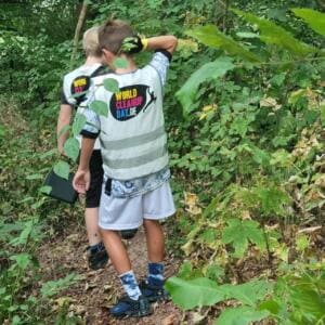 Zwei Jungen in weißen World-Cleanup-Day-Warnwesten gehen mit einem Eimer durch dichtes Gebüsch, von hinten fotografiert.
