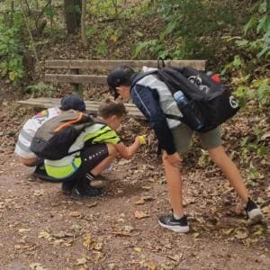 Drei Jungen mit Rucksäcken und Warnwesten bücken sich vor einer Holzbank auf einem laubbedeckten Waldweg und suchen nach Müll.