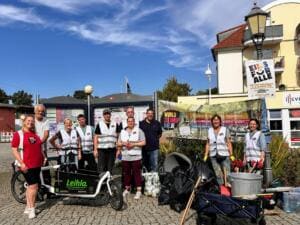 Eine Gruppe Erwachsener in Warnwesten steht auf einem gepflasterten Platz um ein Lastenrad, daneben Bollerwagen mit Säcken, Greifzangen und ein World-Cleanup-Day-Banner.