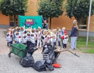 Eine Kindergartengruppe in World-Cleanup-Day-Westen steht vor einer grünen World-Cleanup-Day-Fahne, davor Bollerwagen und gefüllte Müllsäcke.