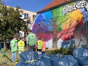 Kinder und ein Erwachsener in Warnwesten stehen vor einer bunten Graffiti-Wand mit der Aufschrift Falkensee, davor viele volle blaue Müllsäcke.