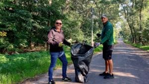 Zwei ältere Männer halten auf einem Waldweg gemeinsam einen schwarzen Müllsack auf, einer mit Greifzange.